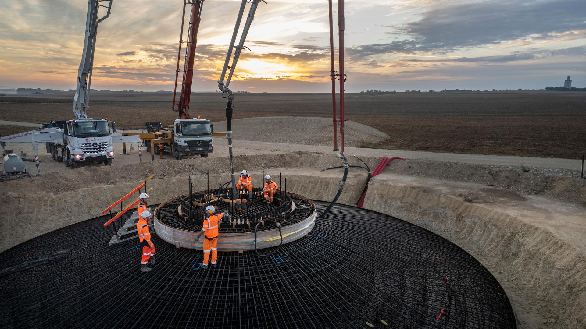 Wind farm construction site showing workers and concrete pumping during turbine foundation installation