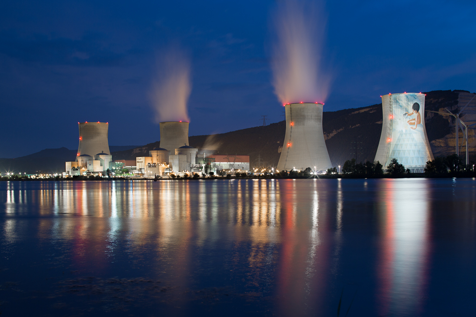 Nuclear power plant at night with illuminated cooling towers reflected on calm water, showcasing large-scale energy infrastructure