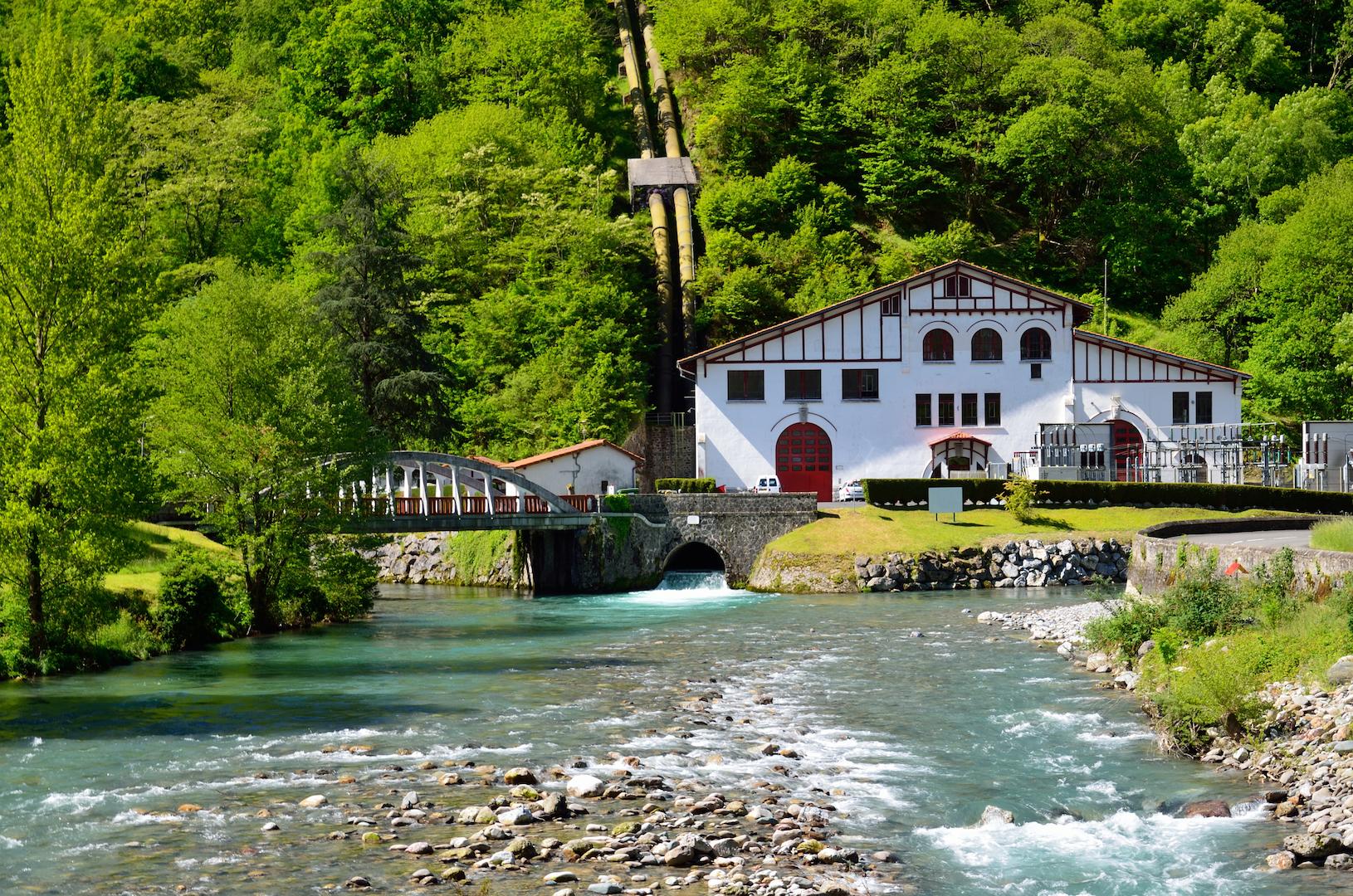 Small hydroelectric compensator power station by a river, featuring a traditional powerhouse building, water channel, and surrounding forest, supporting sustainable local energy generation.