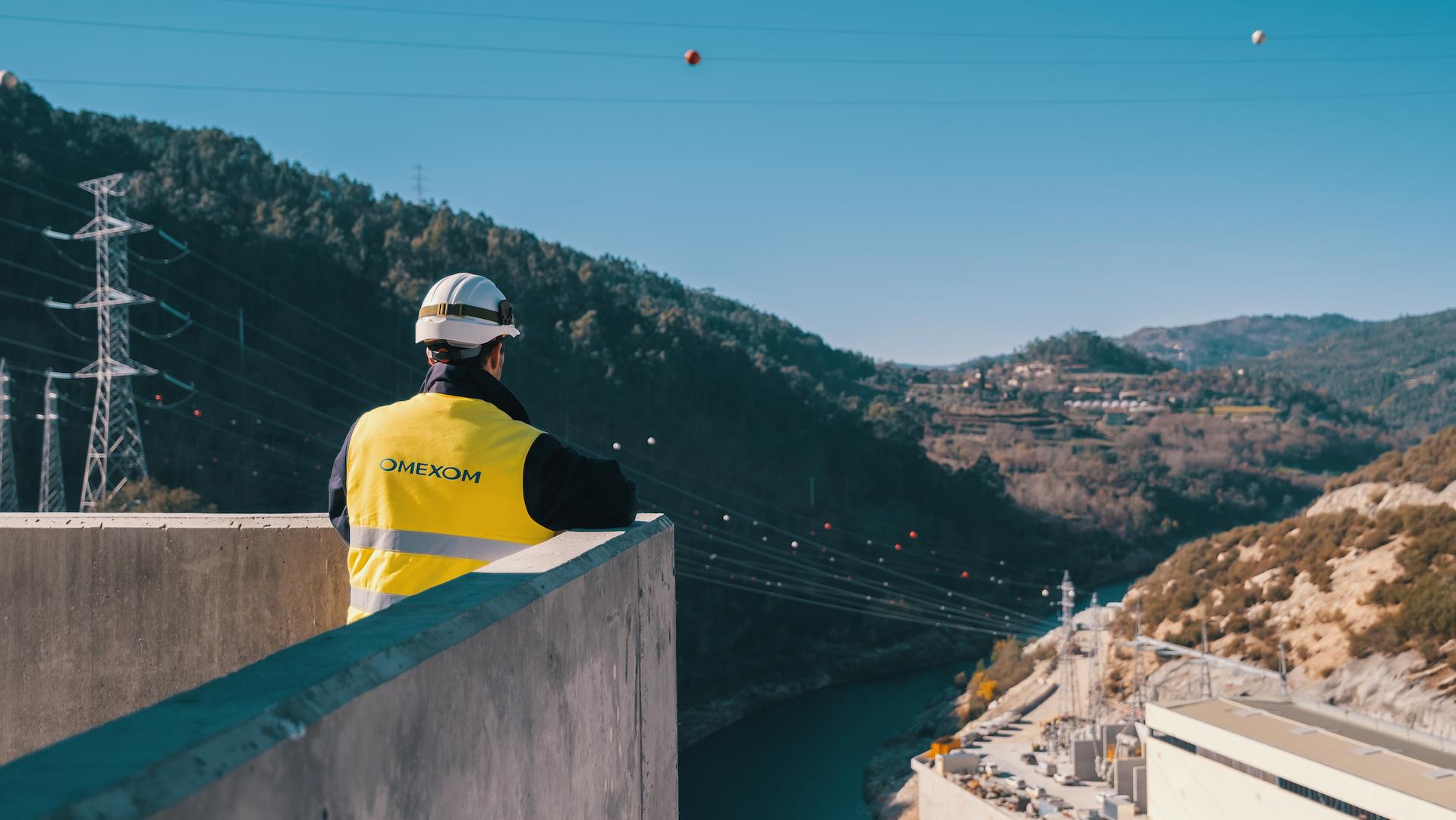 Omexom technician overlooking the Tâmega hydroelectric power station in northern Portugal, with transmission lines and river valley illustrating sustainable energy generation.