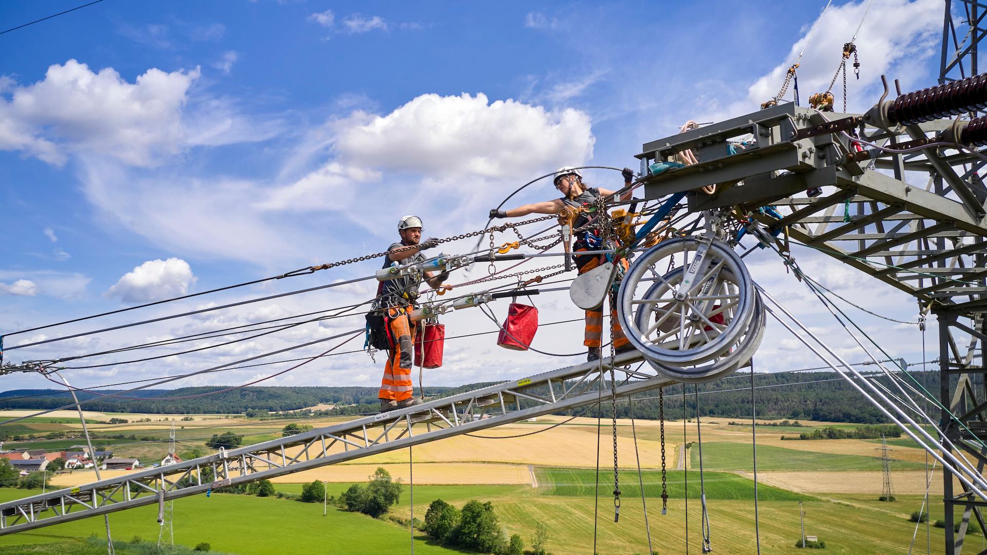 Technicians installing overhead high-voltage transmission conductors on a power line tower in a rural landscape