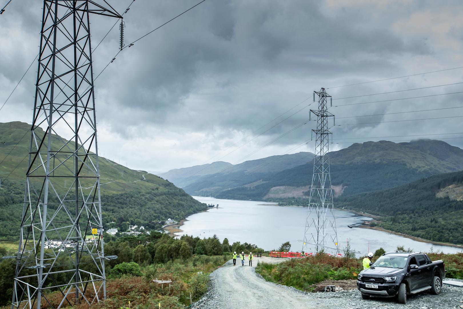 Overhead power transmission pylons crossing a remote valley with a loch, part of a highvoltage electricity network