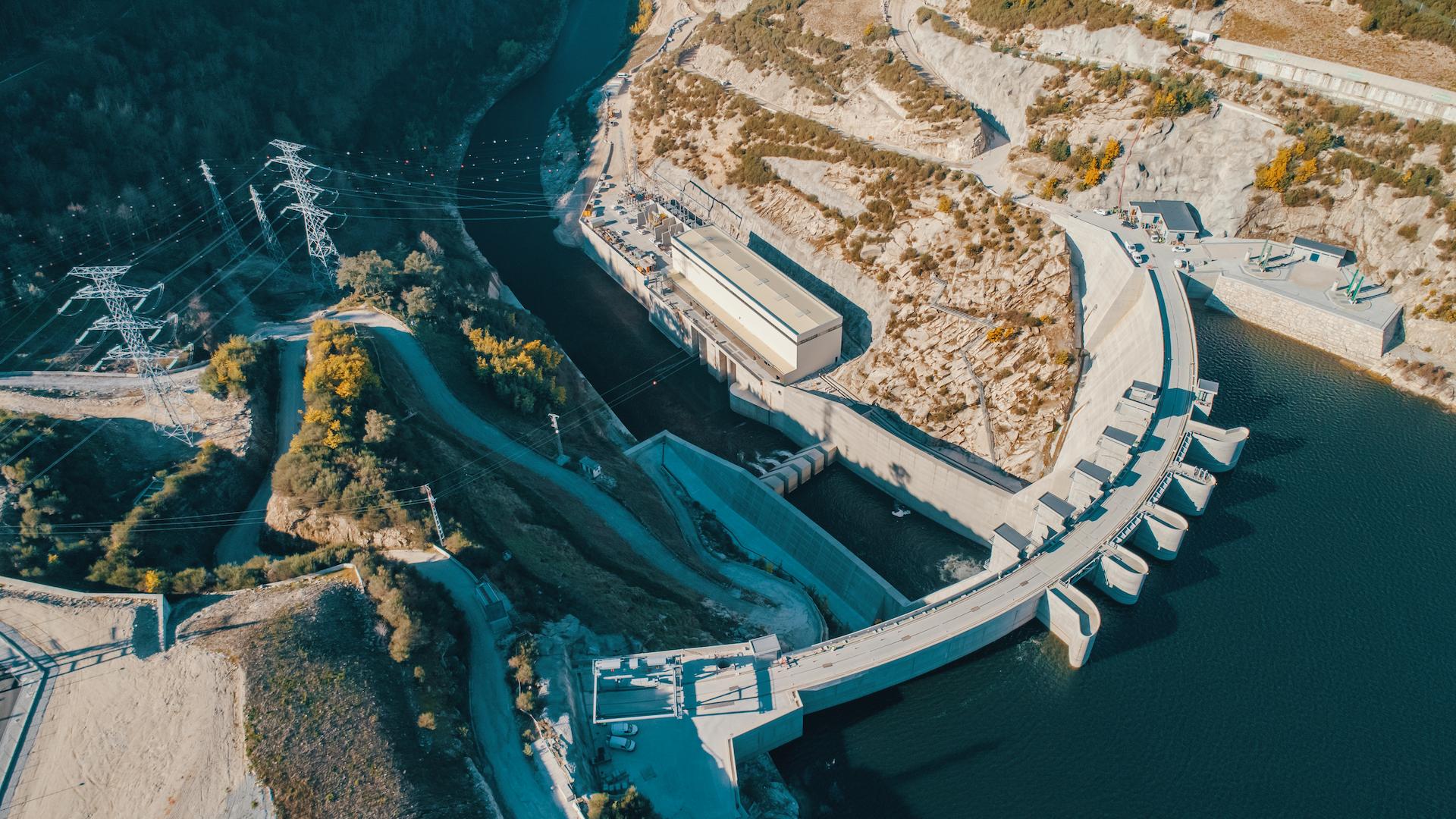 Aerial view of the Tâmega hydroelectric dam in northern Portugal, featuring the curved dam structure, powerhouse, spillways, and high-voltage transmission lines within a mountainous valley.