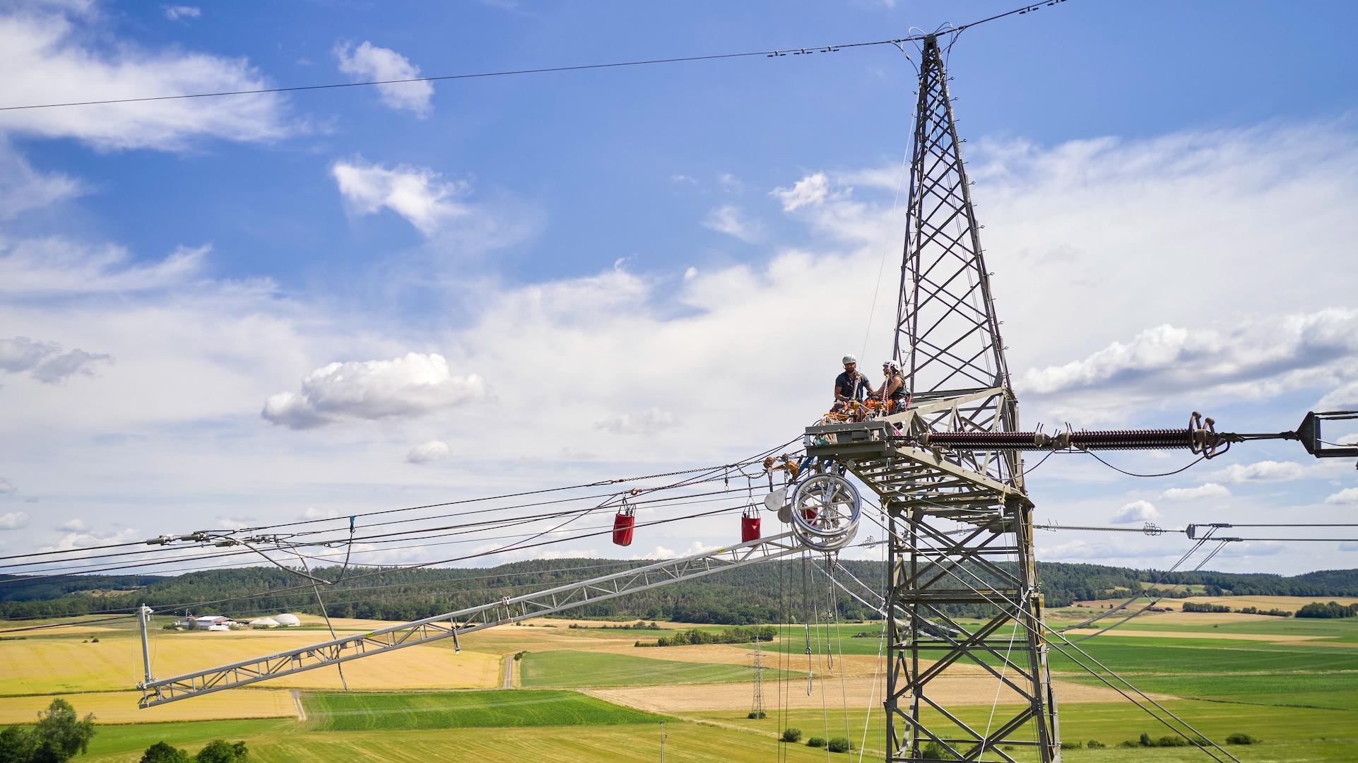 Overhead transmission line technicians working on a highvoltage electricity pylon above farmland, maintaining power grid infrastructure