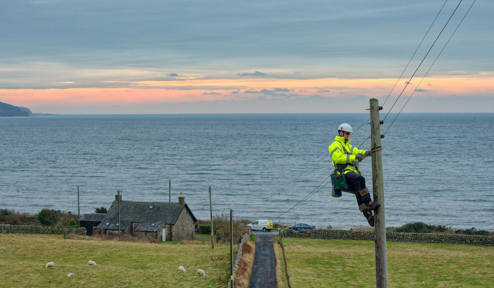 Electricity distribution fitter working on overhead lines from a utility pole, maintaining power supply infrastructure in a coastal rural area