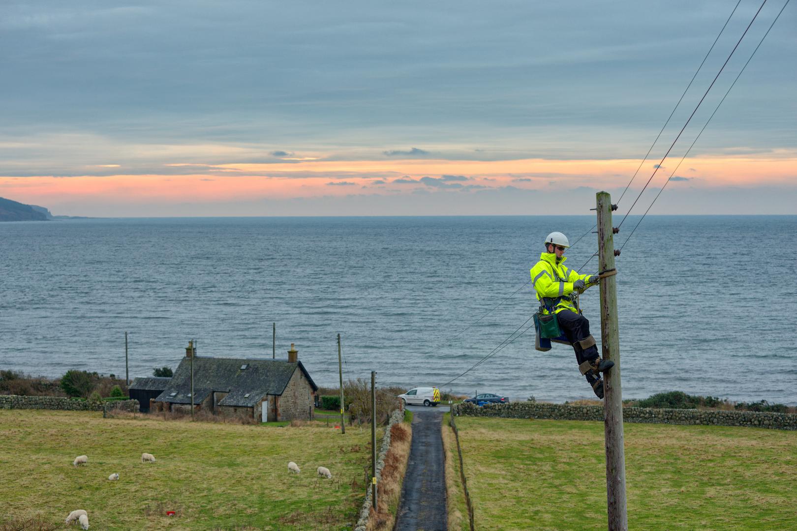 Electricity distribution fitter working on overhead lines from a utility pole, maintaining power supply infrastructure in a coastal rural area