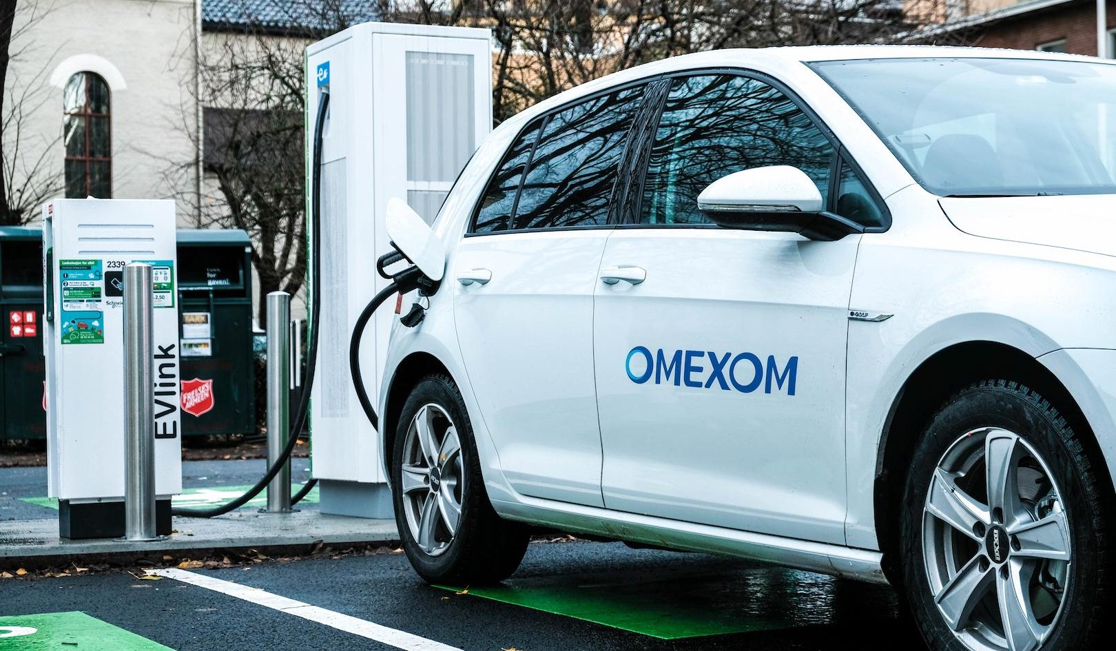 Omexom electric vehicle charging station in an urban environment, with a white EV connected to a public charging point supporting sustainable mobility
