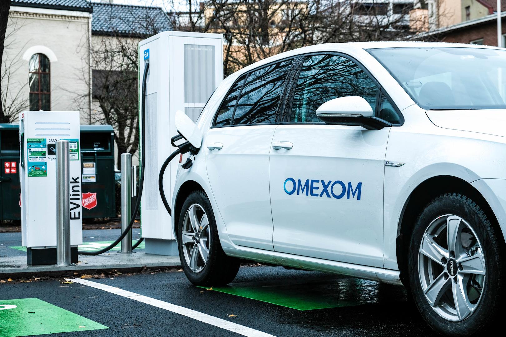 Omexom electric vehicle charging station in an urban environment, with a white EV connected to a public charging point supporting sustainable mobility