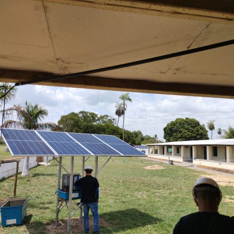Small offgrid solar installation with groundmounted panels powering equipment in a rural area surrounded by buildings and palm trees.