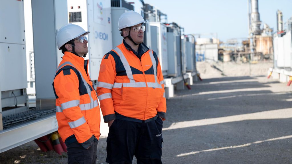 Engineers wearing safety gear standing beside containerized battery storage units at an energy storage facility.