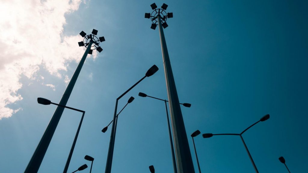 Tall urban street lighting poles viewed from below against a blue sky, illustrating large-scale outdoor lighting infrastructure