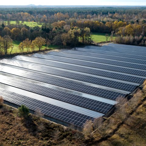 Aerial view of a large solar farm with rows of dark photovoltaic panels reflecting sunlight, set within a rural landscape surrounded by green fields and autumn-colored trees.