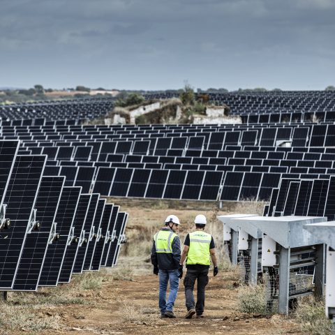 Two technicians wearing high-visibility vests and hard hats walk along a dirt path between rows of solar panels at a large solar farm, with electrical equipment units to the right and rolling fields and a cloudy sky in the background.