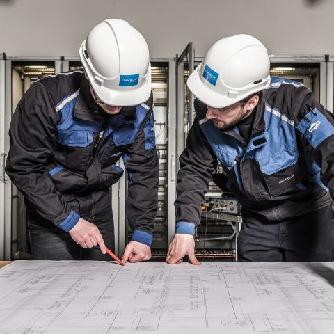 Two workers wearing white hard hats and blue-black work jackets lean over a large technical blueprint spread across a table. One worker points to the drawing with a red pencil while the other follows along. Behind them, rows of open electrical control cabinets and network equipment suggest an industrial or data infrastructure environment.