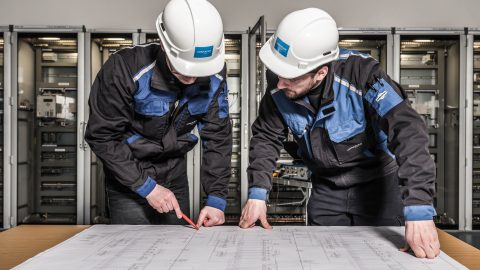 Two workers wearing white hard hats and blue-black work jackets lean over a large technical blueprint spread across a table. One worker points to the drawing with a red pencil while the other follows along. Behind them, rows of open electrical control cabinets and network equipment suggest an industrial or data infrastructure environment.